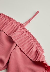 Vaaleanpunainen hihaton vaate, jossa on ohut olkaimet ja röyhelökaulus, valkoista taustaa vasten.