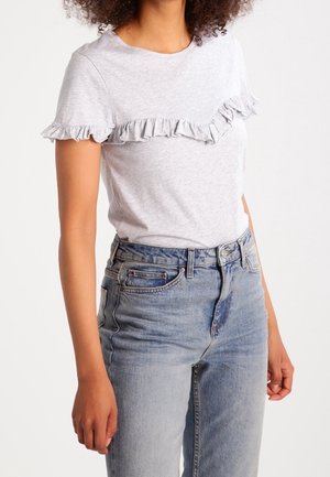 Kvinde iført lysegrå kortærmet top med flæsedetaljer over bryst og ærmer, kombineret med højtaljede blå jeans.