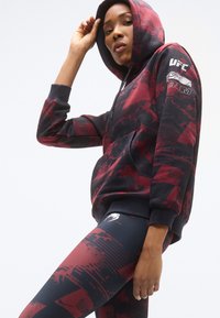 Jopica s kapuco v temno rdeči in črni barvi z abstraktnimi vzorci, ki ima žep vrečko in logotip UFC. Legice v ujemajoči se barvni shemi z grafičnim dizajnom.