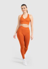 Reggiseno sportivo arancione a coste e leggings a vita alta con design senza cuciture, abbinati a scarpe da ginnastica bianche, su uno sfondo grigio.