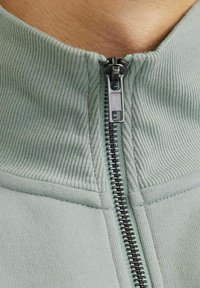 Green zip-up jacket featuring a ribbed collar, textured fabric, and a contrasting black zipper along the front.