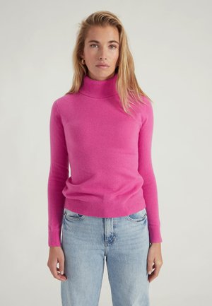 Maglione aderente rosa a collo alto realizzato in materiale morbido, caratterizzato da maniche lunghe e orlo a costine, abbinato a jeans azzurri chiari.