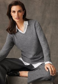 Szary sweter w kształcie litery V z miękkiego materiału w luźnym kroju, nałożony na białą koszulę. Model nosi eleganckie szare spodnie. Proste okrągłe kolczyki.