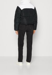 Osoba v černé a bílé péřové bundě, černých džínách a bílých teniskách stojí proti jednobarevnému světlému pozadí, viděná zezadu.