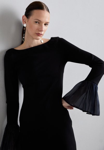 Black dress with long sleeves, sheer navy bell cuffs, boat neckline, and a fitted silhouette. Large statement earrings complete the look.