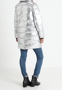 Srebrna pikowana kurtka puchowa z poziomym wzorem pikowania, o długości do połowy uda, z szerokim kołnierzem, zestawiona z niebieskimi dżinsami i czarnymi botkami za kostkę.
