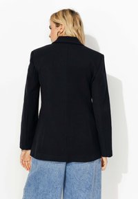 Femme blonde portant des boucles d'oreilles en or, un blazer noir et un jean large bleu clair, de dos contre un fond blanc.