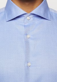 Camicia azzurra chiaro con bottoni, caratterizzata da un motivo a rombi, colletto classico e bottoni bianchi su un tessuto testurizzato.
