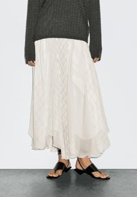 Hellbeige geschichteter Rock mit Diamantmuster, kombiniert mit einem dunkelgrauen Strickpullover und schwarzen flachen Sandalen. Glatter, fließender Stoff.