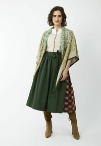 Tenue traditionnelle comprenant un châle à motifs, un haut crème avec des boutons et une jupe verte. Des bottines marron aux chevilles complètent le look.