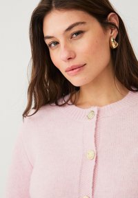Roze gebreide cardigan met een ronde hals, voorzien van gouden knopen en een zachte textuur. Close-up van de halslijn en de knopen.
