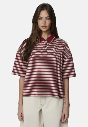 Jeune femme aux longs cheveux bruns portant un polo ample à rayures bordeaux et blanches et un pantalon clair, debout devant un fond uni.
