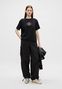 Zwarte katoenen t-shirt met een logo, gecombineerd met losse zwarte cargobroek en zilveren sneakers. Model houdt een zwart leren jasje vast.