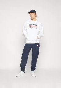 Fehér kapucnis sweatshirt "TOMMY" logóval, tengerészkék melegítőnadrág és fehér sportcipő. A melegítőnadrágon egy kis piros és kék logópárna található. Fesztelen öltözet.