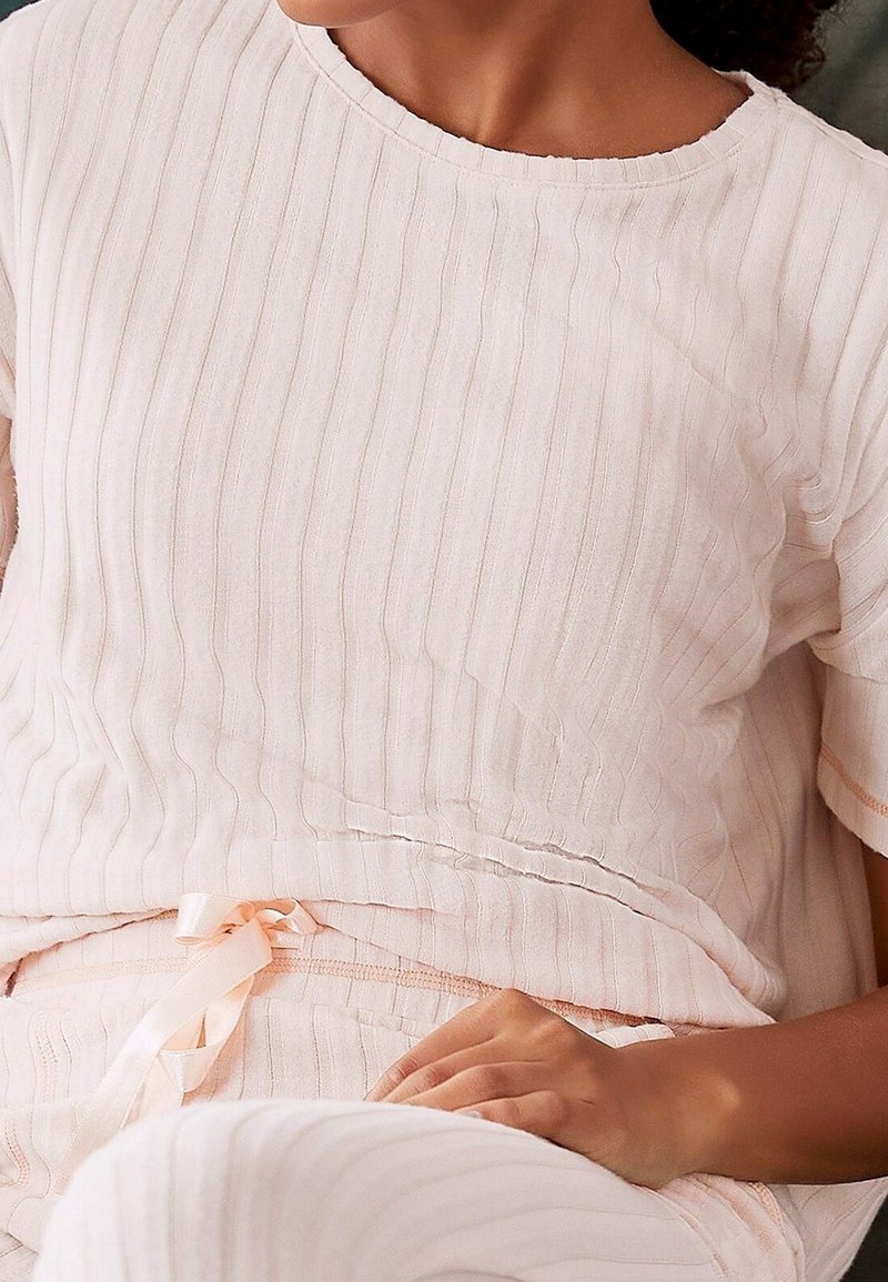Personne portant un haut à manches courtes côtelé rose pâle et un pantalon assorti avec un cordon de serrage en satin, assise avec une main posée sur ses genoux.