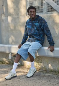 Sort og blå mønstret Nike hættetrøje med logo, lyseblå denimshorts og lyse sneakers med mørke såler, siddende på en bænk.