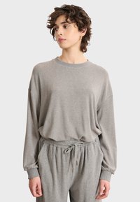 Giovane con i capelli corti e ricci che indossa un ampio maglione grigio chiaro e pantaloni coordinati con coulisse, mentre guarda di lato.