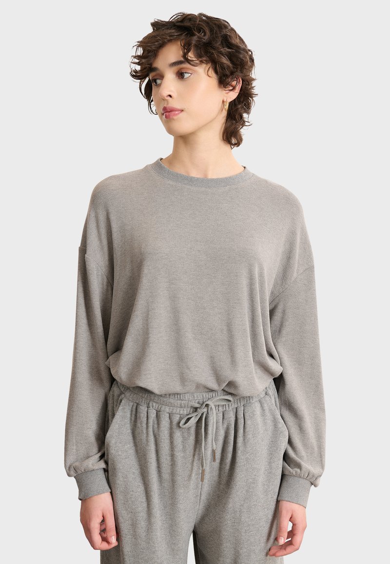 Giovane con i capelli corti e ricci che indossa un ampio maglione grigio chiaro e pantaloni coordinati con coulisse, mentre guarda di lato.