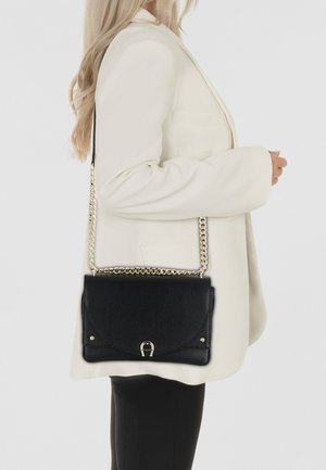 Schwarz strukturiertes Lederhandtasche mit einer goldenen Kettenriemen, mit einer Klappenverschlus und einem metallischen Akzent. Im Crossbody-Stil getragen.