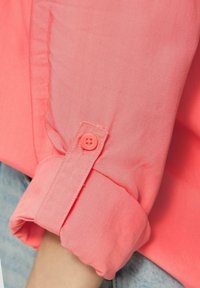 Camicia a manica lunga di un luminoso corallo, con polsini a bottone. Il tessuto è liscio con una leggera lucentezza e i polsini sono arrotolati che rivelano un denim sottostante.