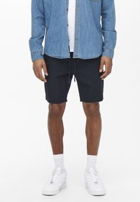 Jeanshemd, blau, mit Knöpfen und einer Brusttasche. Marineblaue Shorts, knielang, mit Kordelzug. Weiße Sneaker mit perforierten Einsätzen.