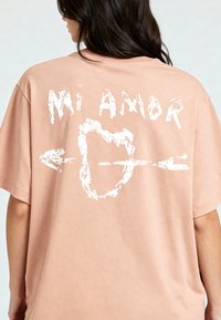 T-shirt rose surdimensionnée en tissu doux, présentant un graphique blanc au dos avec le texte "Mi Amor" et un cœur avec une flèche.