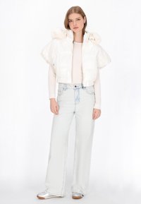 Krótka kurtka puchowa z połyskującego białego materiału z kapturem ozdobionym futrzanymi akcentami, zestawiona z jasnoniebieskimi dżinsami o szerokich nogawkach i srebrnymi trampkami.