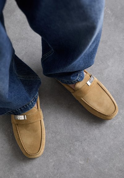 Moccasini din suede bej cu o textură netedă, accente din metal argintiu dreptunghiular, și detalii vizibile de cusături, asortați cu blugi din denim albastru.