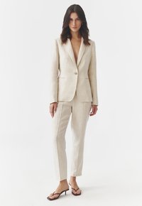Beige lenvászon öltöny, kiflivel ellátott blézerrel és slim-fit nadrággal. Az öltözék diszkrét textúrával és minimalista dizájnnal rendelkezik, vékony sarkú szandálokkal párosítva.