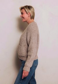 Beiger Strickpullover mit strukturierter Oberfläche, ausgestattet mit einer Fronttasche, getragen über blauen Jeans, seitlich betrachtet vor hellem Hintergrund.