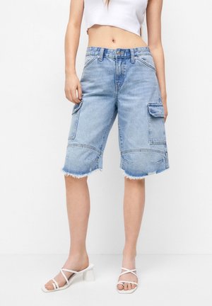 Femme portant un short cargo en jean bleu clair avec des bords effilochés et des sandales blanches à talons avec des lanières, debout devant un fond blanc.