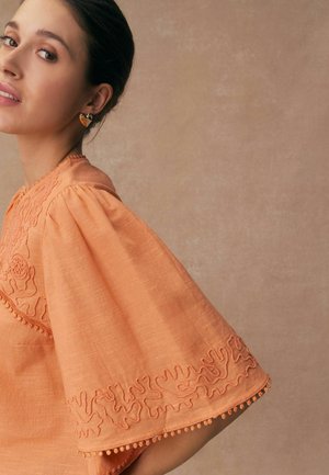 Woman wearing an orange blouse with wide, embroidered sleeves and heart-shaped earrings against a beige background.