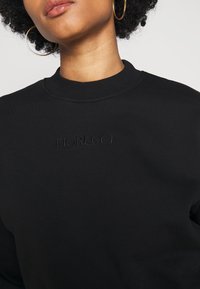 Gros plan d'une personne portant de grandes boucles d'oreilles créoles dorées et un sweat-shirt noir avec le logo brodé "FIORUCCI" sur la poitrine.