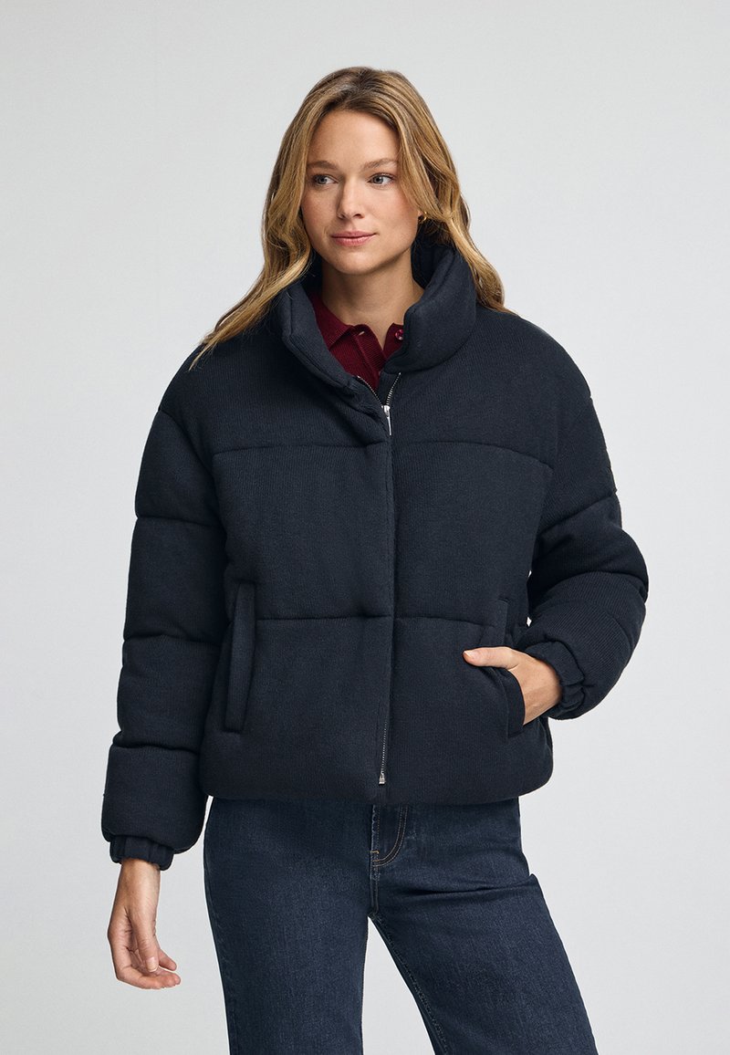 Mujer con una chaqueta acolchada negra de tipo puffer con un cuello alto, una mano en el bolsillo, combinada con jeans oscuros y una blusa burdeos debajo.