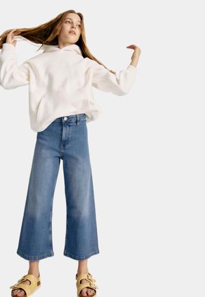 Biała bluza z polaru z kieszenią z przodu, zestawiona z szerokimi niebieskimi dżinsami. Model nosi beżowe sandały z klamrami.