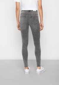 Person som bär åtsittande grå jeans och vita sneakers, står med ryggen mot kameran mot en enkel ljus bakgrund.