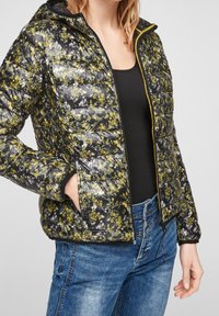 Blomstret pufferjakke med et sort og gult mønster, letvægts, lynlåslukning foran, hætte, sidelommer og ribbede manchetter, parret med blå jeans.