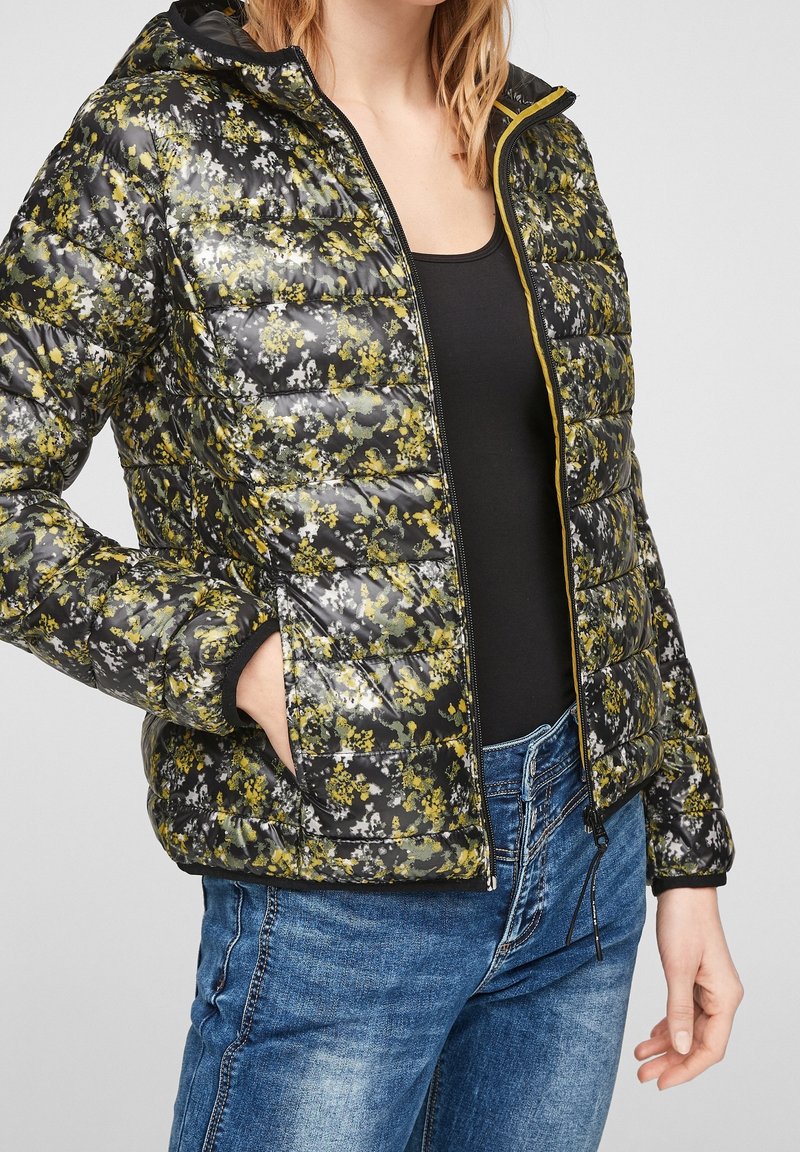 Blomstret pufferjakke med et sort og gult mønster, letvægts, lynlåslukning foran, hætte, sidelommer og ribbede manchetter, parret med blå jeans.