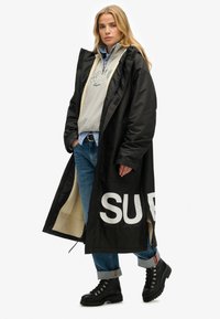 Abrigo negro largo con letras blancas "SUP", forrado con forro polar beige. Lleva puesto sobre un sudadera gris, una camisa azul y jeans, combinado con botas negras.