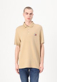 Beige polo-skjorte med krage, korte ermer og tre knapper. Har et lite emblem med røde, hvite og blå detaljer.