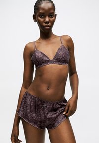 Γυναίκα με σκούρο δέρμα και πλεγμένα μαλλιά, φοράει μωβ σουτιέν με ζωικά σχέδια και αντίστοιχο σορτς, στέκεται μπροστά σε έναν απλό λευκό φόντο.