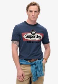 Camiseta de algodón azul marino con cuello redondo y ribetes en negro en contraste. Presenta el logo de "Superdry" en blanco y detalles en rojo. Se lleva con pantalones caqui.