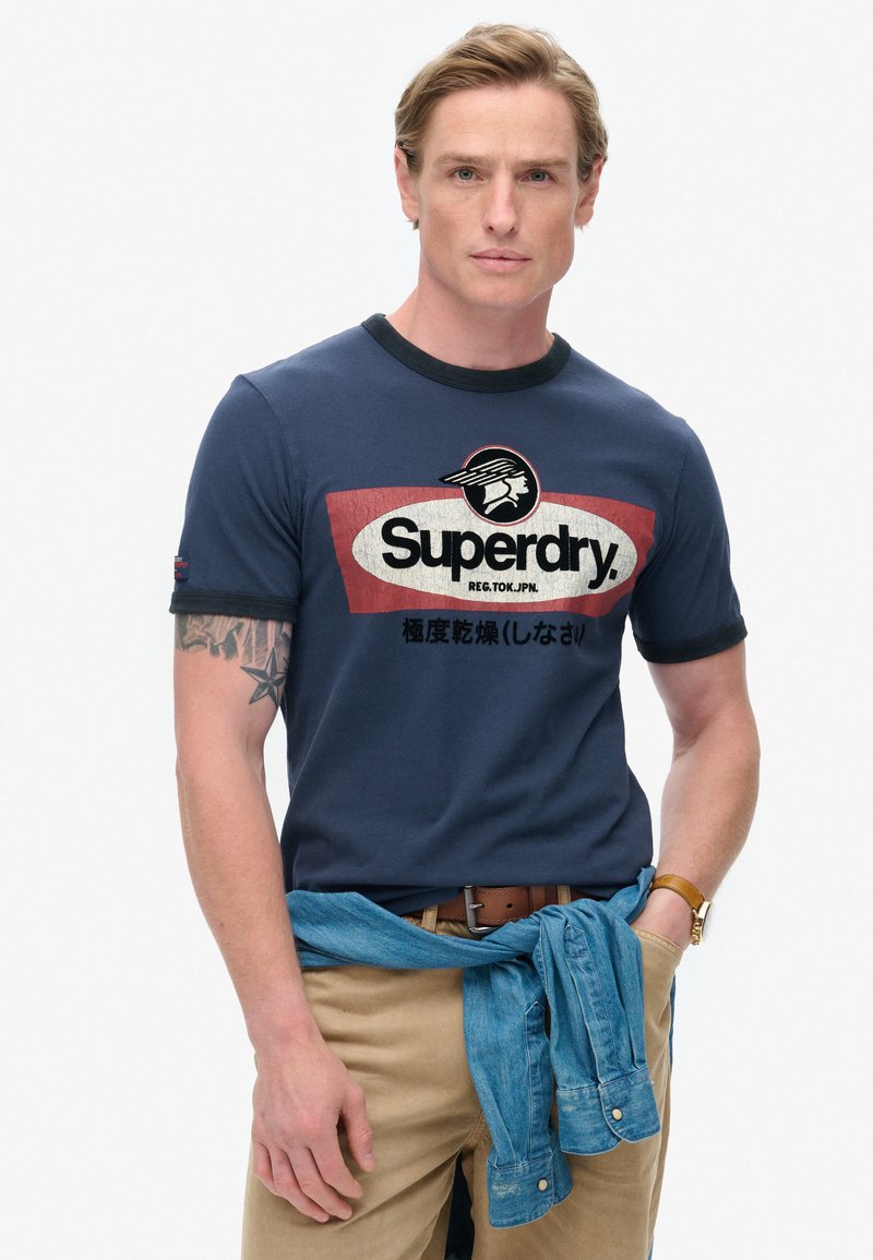 Camiseta de algodón azul marino con cuello redondo y ribetes en negro en contraste. Presenta el logo de "Superdry" en blanco y detalles en rojo. Se lleva con pantalones caqui.