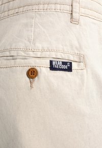 Tejido beige con un bolsillo que presenta un pequeño botón y una etiqueta azul marino que dice "VISTE EL CÓDIGO".