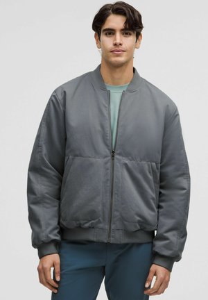 Nuori mies, joka pukeutunut harmaaseen vetoketjulliseen bomber-takkiin vaaleanvihreän paidan ja sinisten housujen kanssa, seisoo yksinkertaisen vaalean taustan edessä.