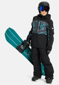 Schwarze Snowboardjacke mit türkisfarbenen Akzenten, kombiniert mit schwarzen Hosen. Hält ein türkisfarbenes Snowboard mit weißen Streifen und schwarzen Bindungen. Schwarzer Helm und Goggles.