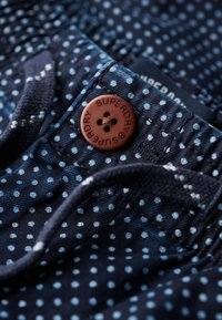 Tissu bleu marine à pois blancs, avec un bouton marron portant l'inscription "SUPERDRY" en relief et des cordons pour réglage.