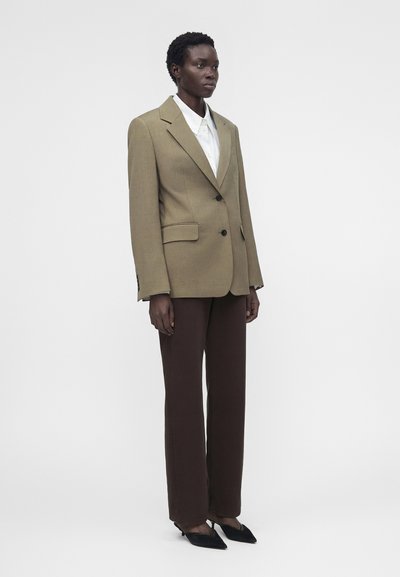 Άτομο στέκεται φορώντας ανοιχτό καφέ σακάκι πάνω από άσπρο πουκάμισο, σκούρο καφέ παντελόνι και μαύρα μυτερά παπούτσια σε απλό φόντο.