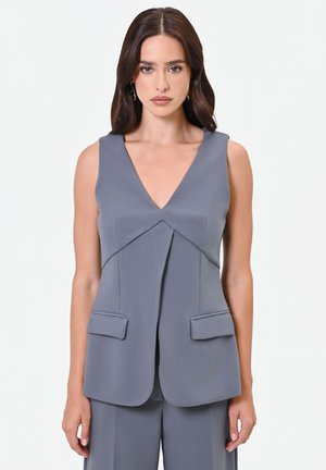 Vrouw met lang bruin haar, dragend een mouwloos grijs op maat gemaakt vest met voorflapzakken en een bijpassende grijze broek tegen een effen achtergrond.