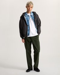 Sweatshirt à capuche gris foncé superposé sur une veste en denim bleu clair, un t-shirt blanc, un pantalon vert et des baskets noires.